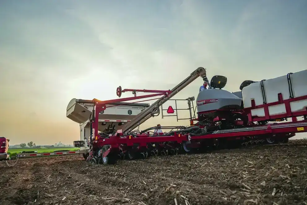 A large, red Case IH agricultural planter is in a field under a cloudy sky, surrounded by tilled soil. The scene conveys productivity and modern farming.