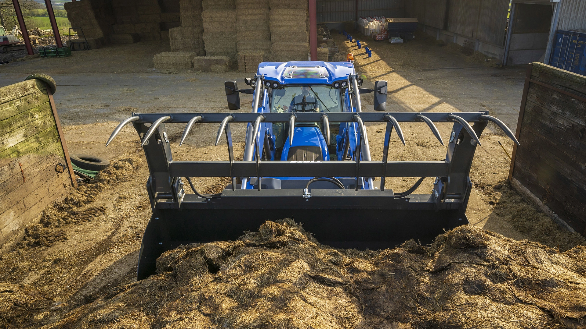 Front loader attachment on a New Holland tractor maneuvering in a barnyard.