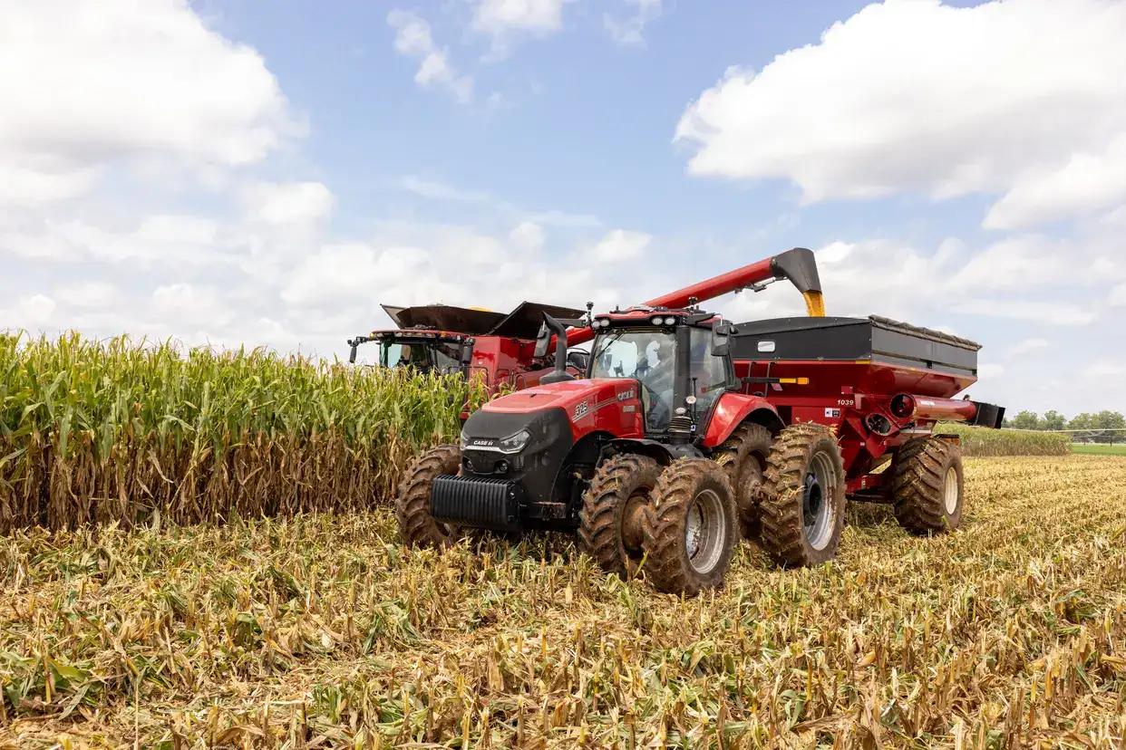 A Case IH tractor and combine harvester work side by side in a cornfield under a partly cloudy sky