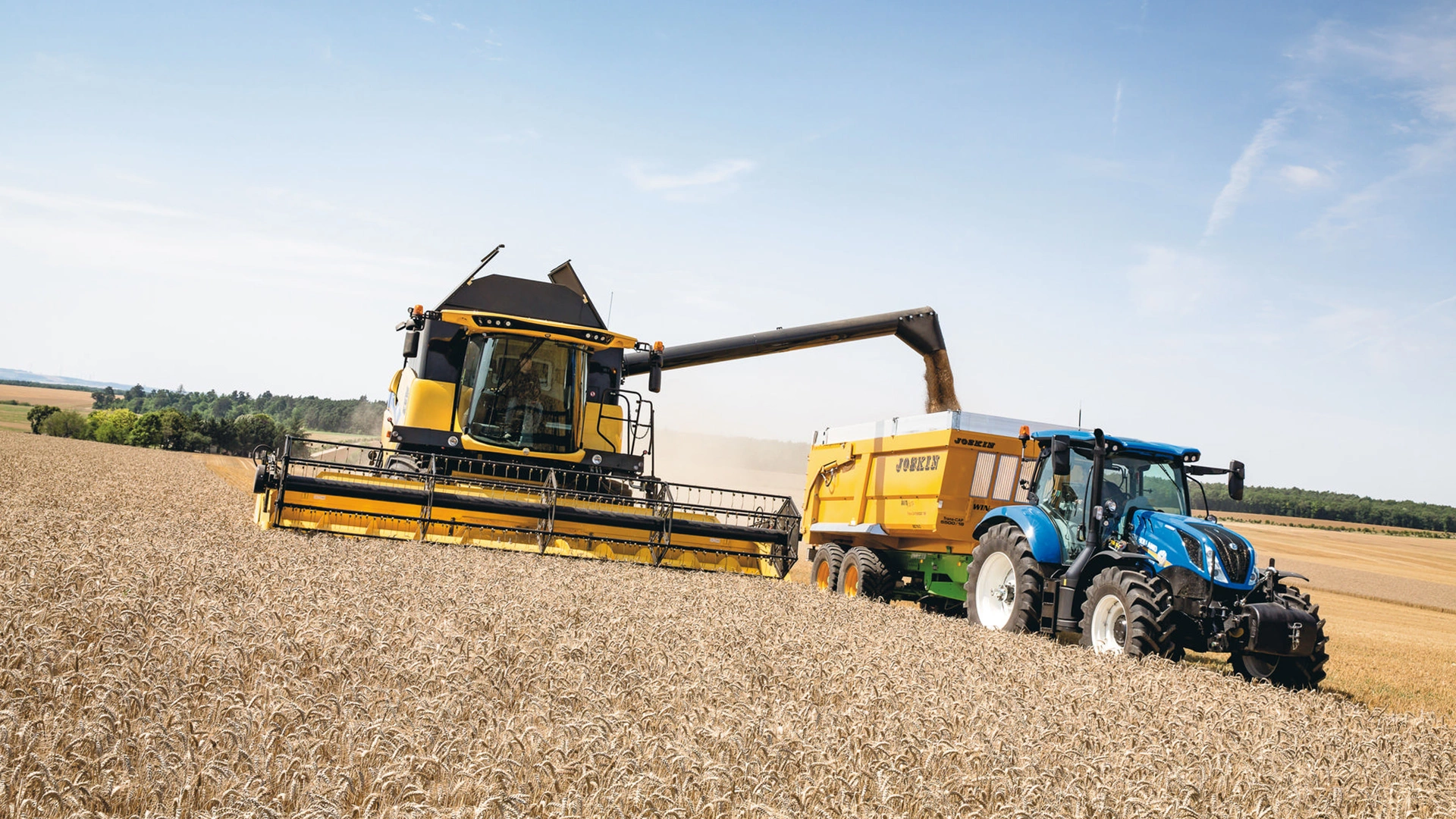 New Holland CH Combine Harvester working in a field, unloading grain into a trailer pulled by a blue tractor, under a clear sky.