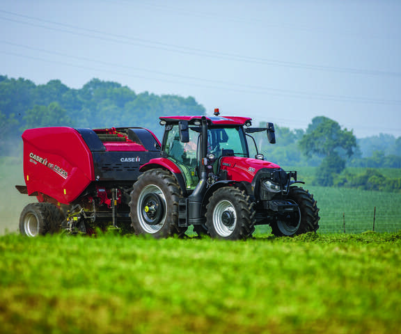 A Case IH tractor with a hay baler attachment operates in a lush, green field under a clear sky. 