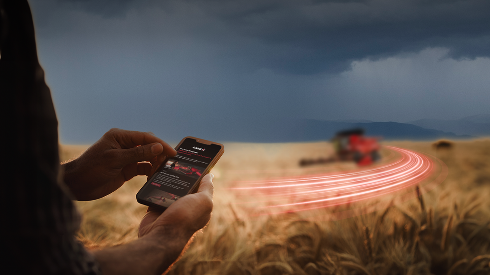Close-up of a farmer's hands holding a smartphone with a Case IH app on the screen in a wheat field, with a Case IH combine harvester and red light trails in the background under a stormy sky