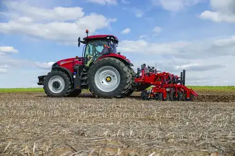 Case IH tractor with a large tillage implement works across a harvested field beneath a bright blue, cloud-dotted sky, conveying rural productivity.