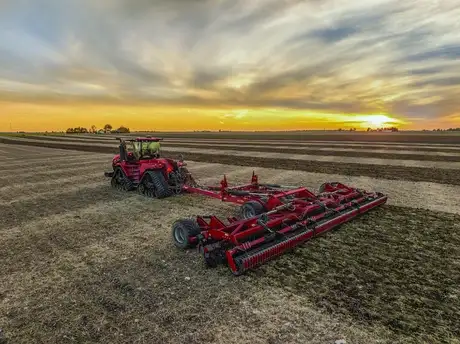 A red Case IH tractor pulls a wide red cultivator across a harvested field at sunset.