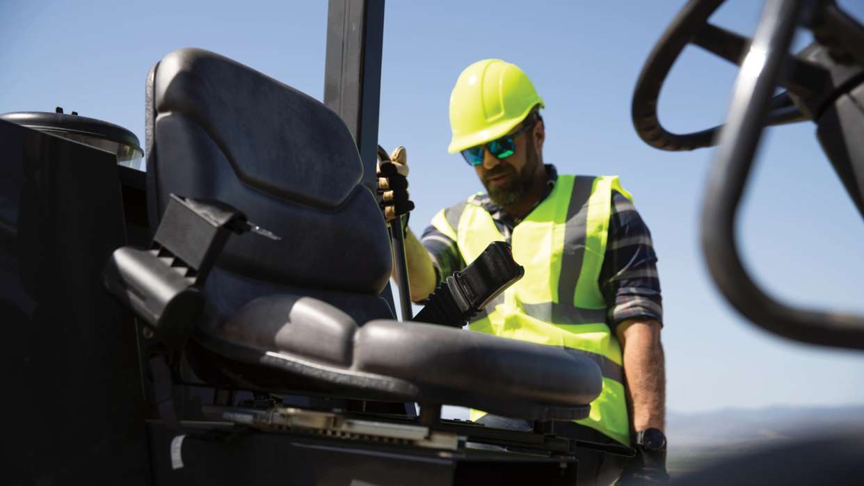 Man in yellow hard hat and vest next to seat of construction equipment.