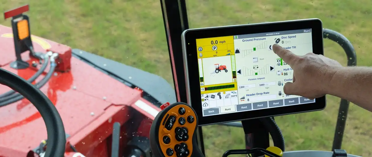 A Case IH tractor operator taps a touchscreen control panel beside an orange joystick; red agricultural mowing equipment is visible outside the cab.