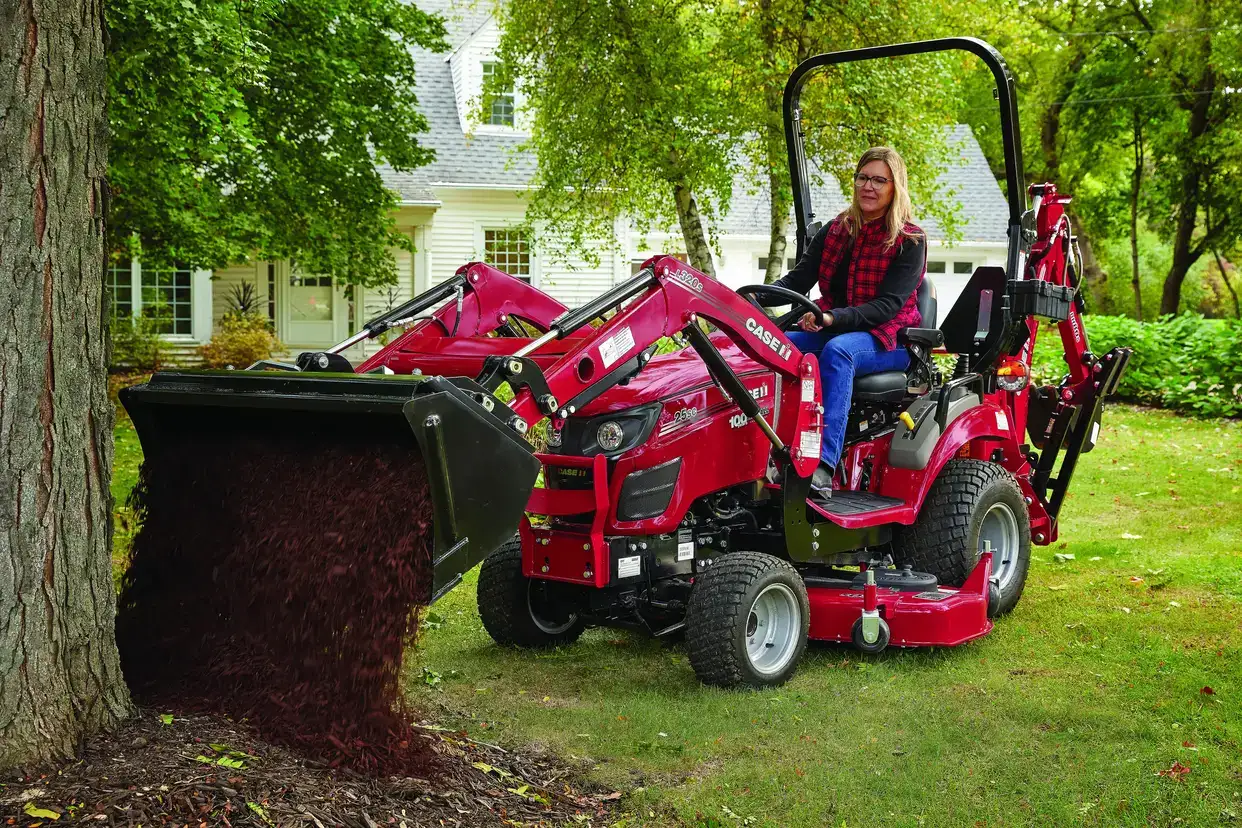 A woman operates a Case IH subcompact tractor, dumping mulch beside a large tree in a garden. 
