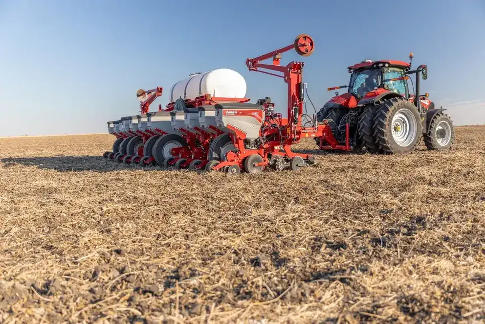 A red Case IH tractor pulls a modern planter across a dry, flat field under a clear blue sky, highlighting agricultural technology and rural landscape.