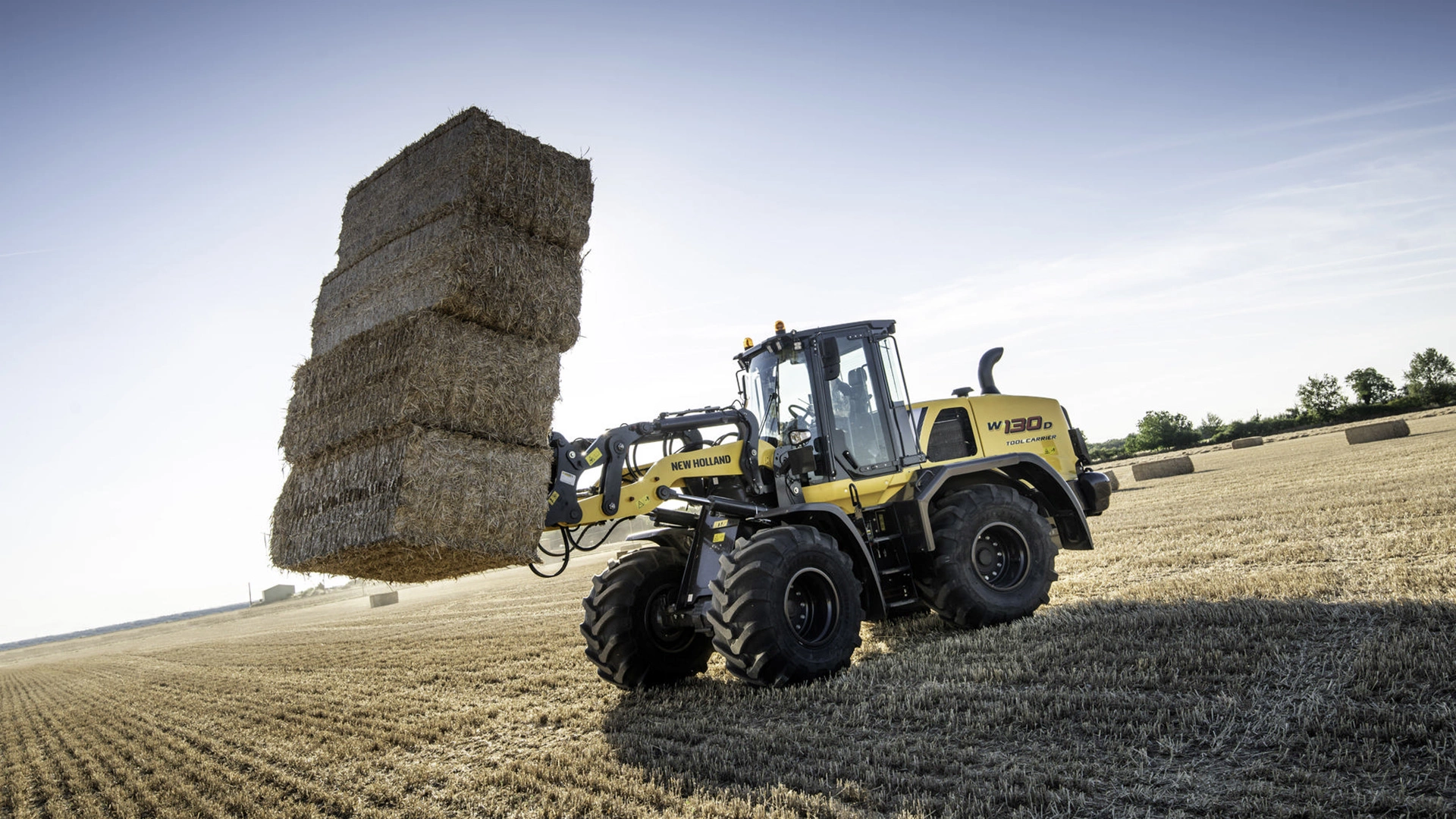 New Holland W130D wheel loader efficiently transporting a towering stack of hay bales on a sunny day in the field.