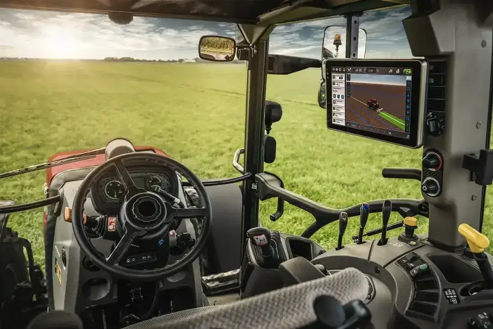 Case IH tractor cab with steering wheel, touchscreen GPS display, and controls overlooking a sunlit green field; advanced agricultural technology.