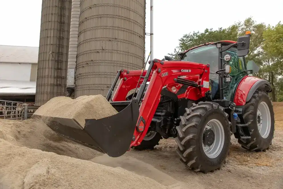 A Case IH tractor with a front loader is scooping sand near large silos on a farm.