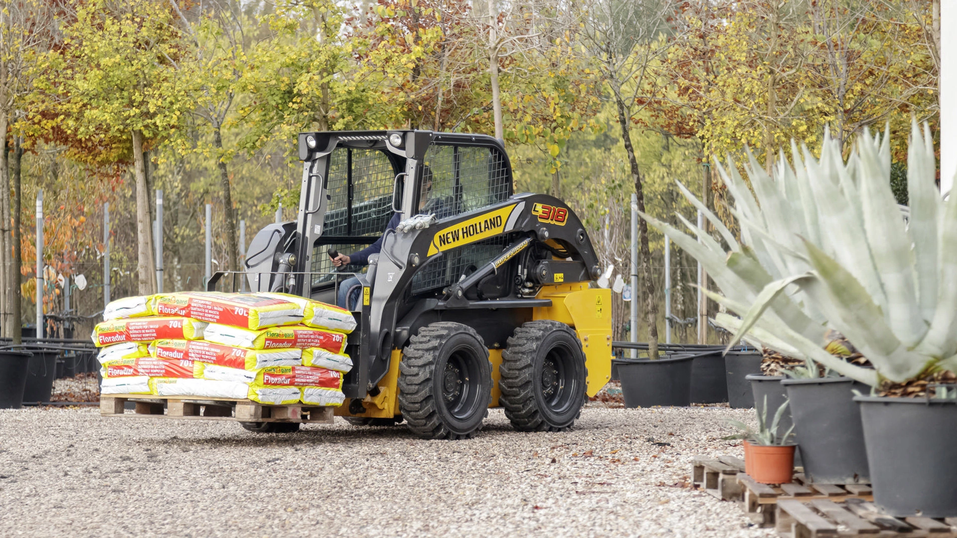 A New Holland skid steer loader in a garden setting, lifting a pallet of soil bags. 
