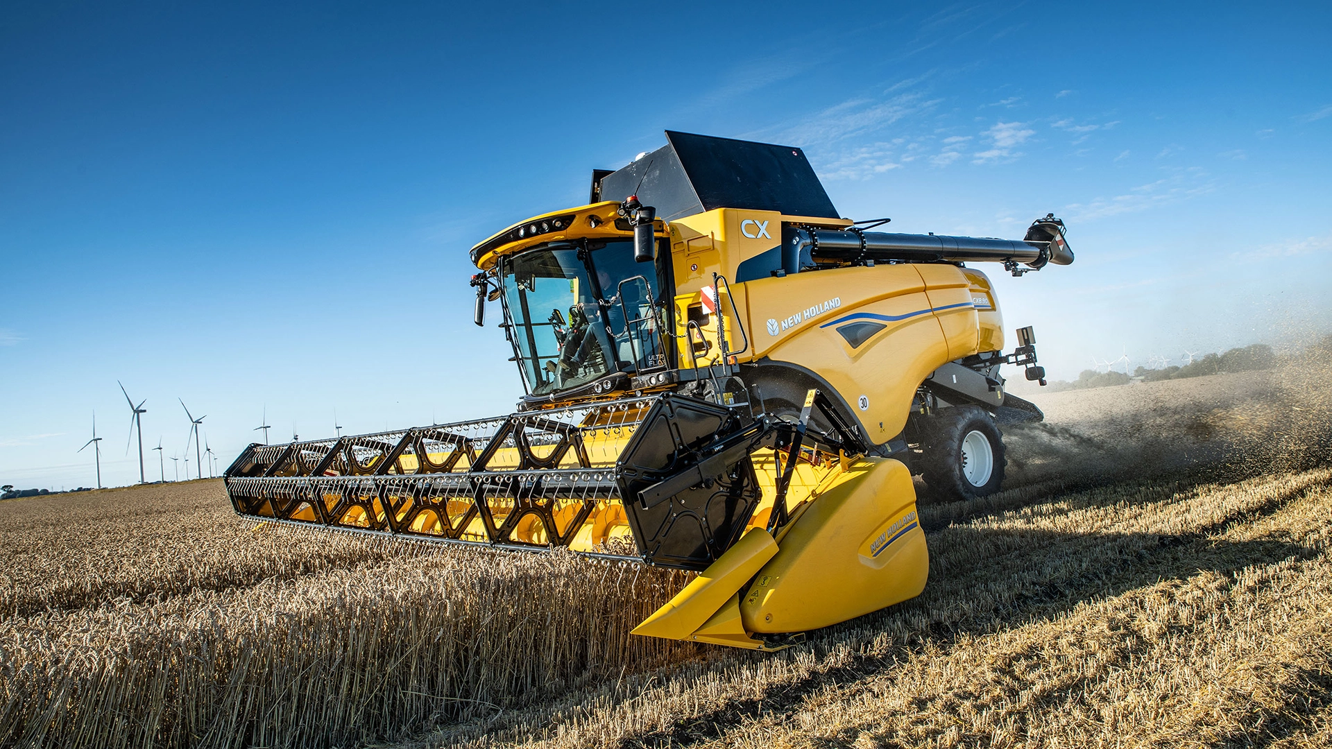 New Holland CX7 & CX8 harvesting golden crops, clear sky with distant wind turbines.