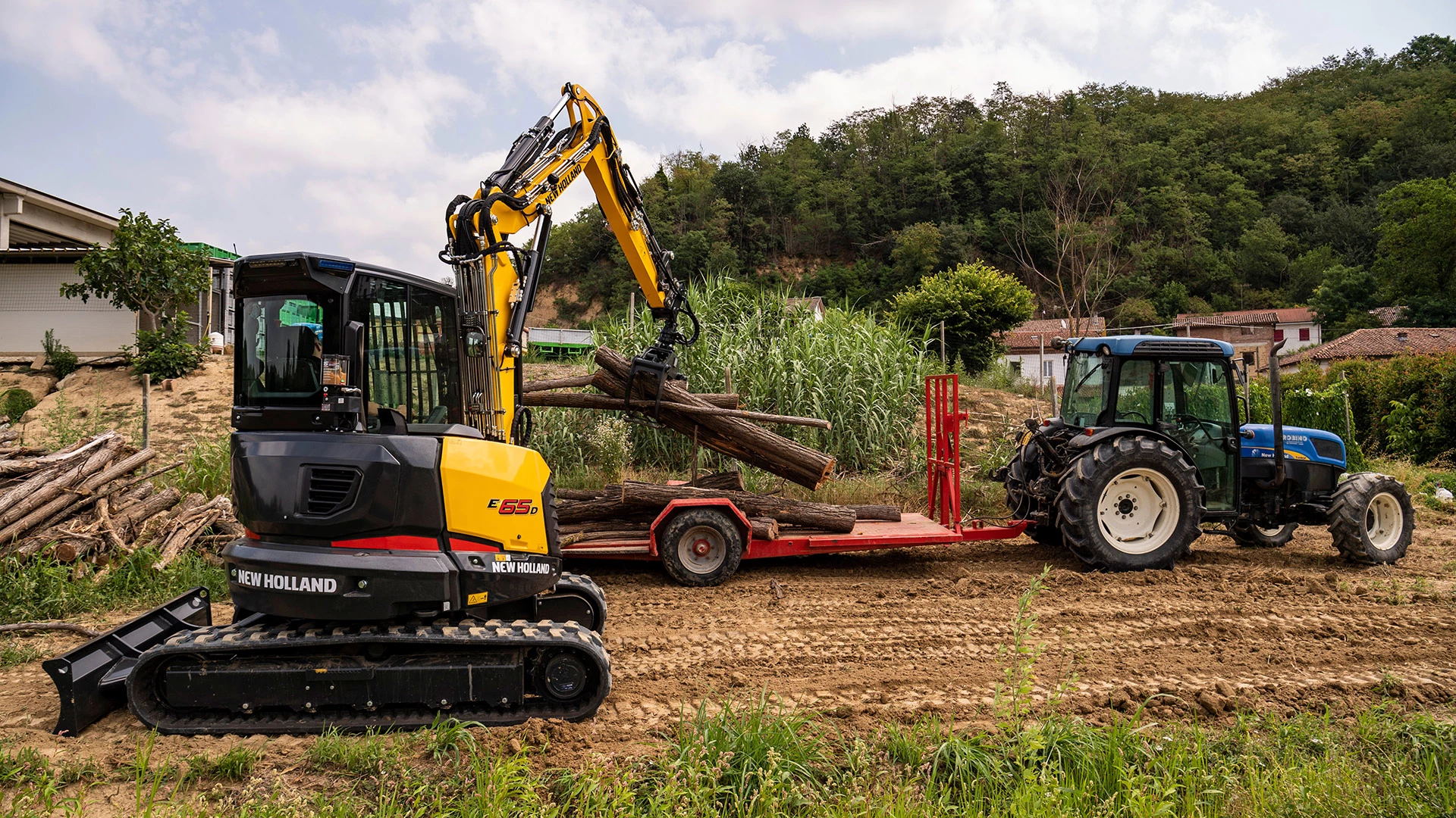 New Holland E55D mini crawler lifting logs, with New Holland tractor & trailer, amidst rural farmland setting.