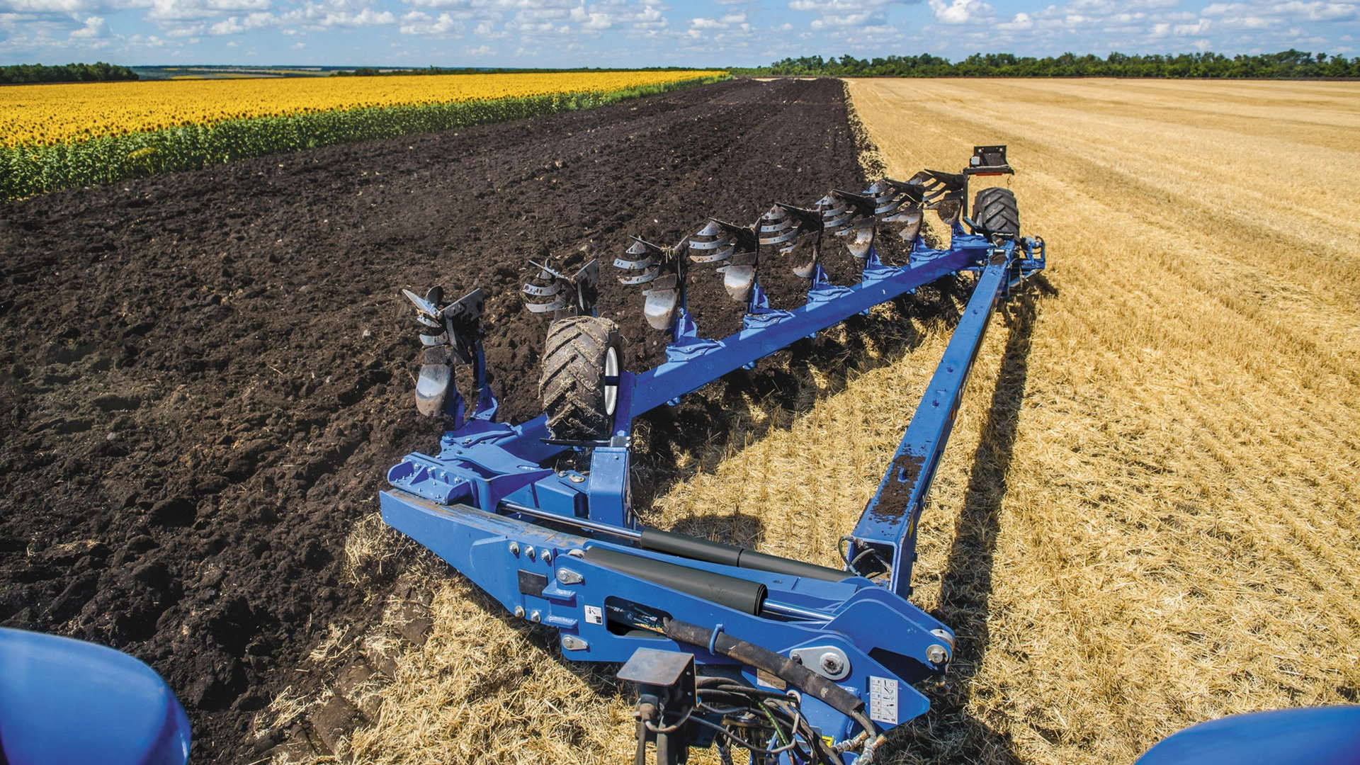 Close-up of an 8-furrow reversible plough at work beside a sunflower field.