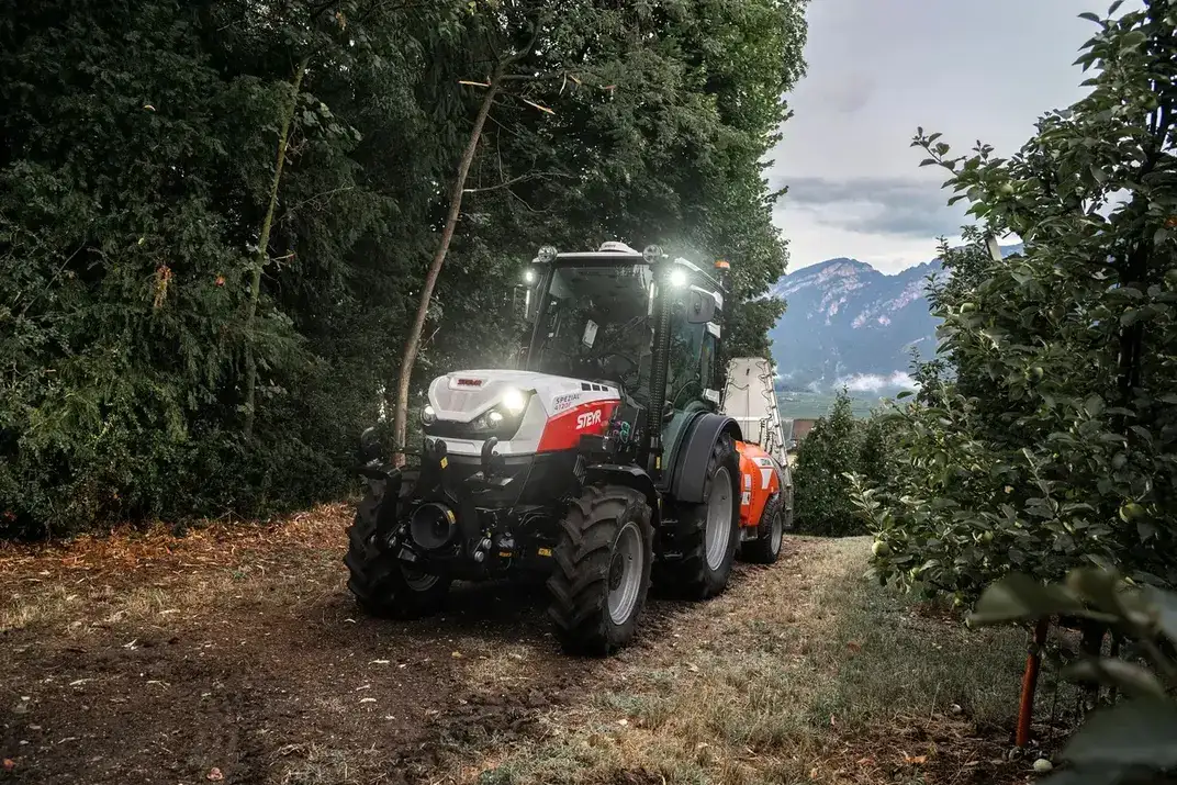STEYR Spezial Obstbautraktor mit orangefarbenem Sprühgerät bei der Arbeit in einer Apfelanlage in der Dämmerung, Scheinwerfer an