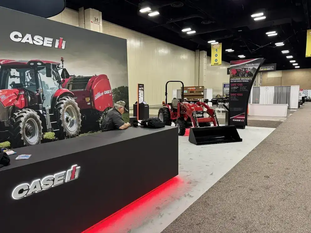 Trade show booth featuring Case IH farm equipment. A large backdrop shows a red tractor. A man sits at a counter with displays and promotional materials, conveying a professional atmosphere.