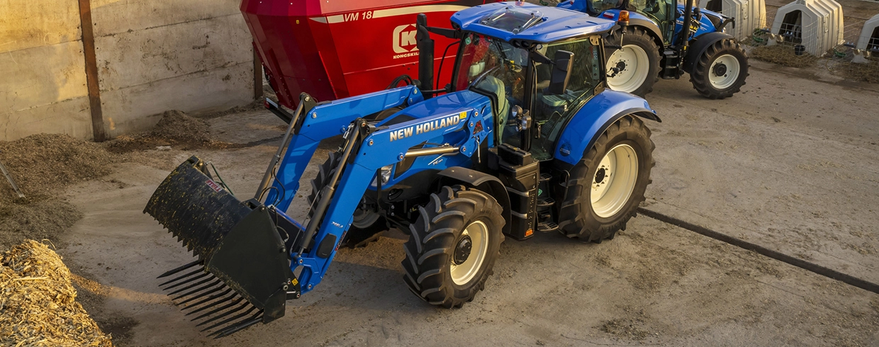 New Holland tractor with front loader bucket working near a feed mixer at a farm.