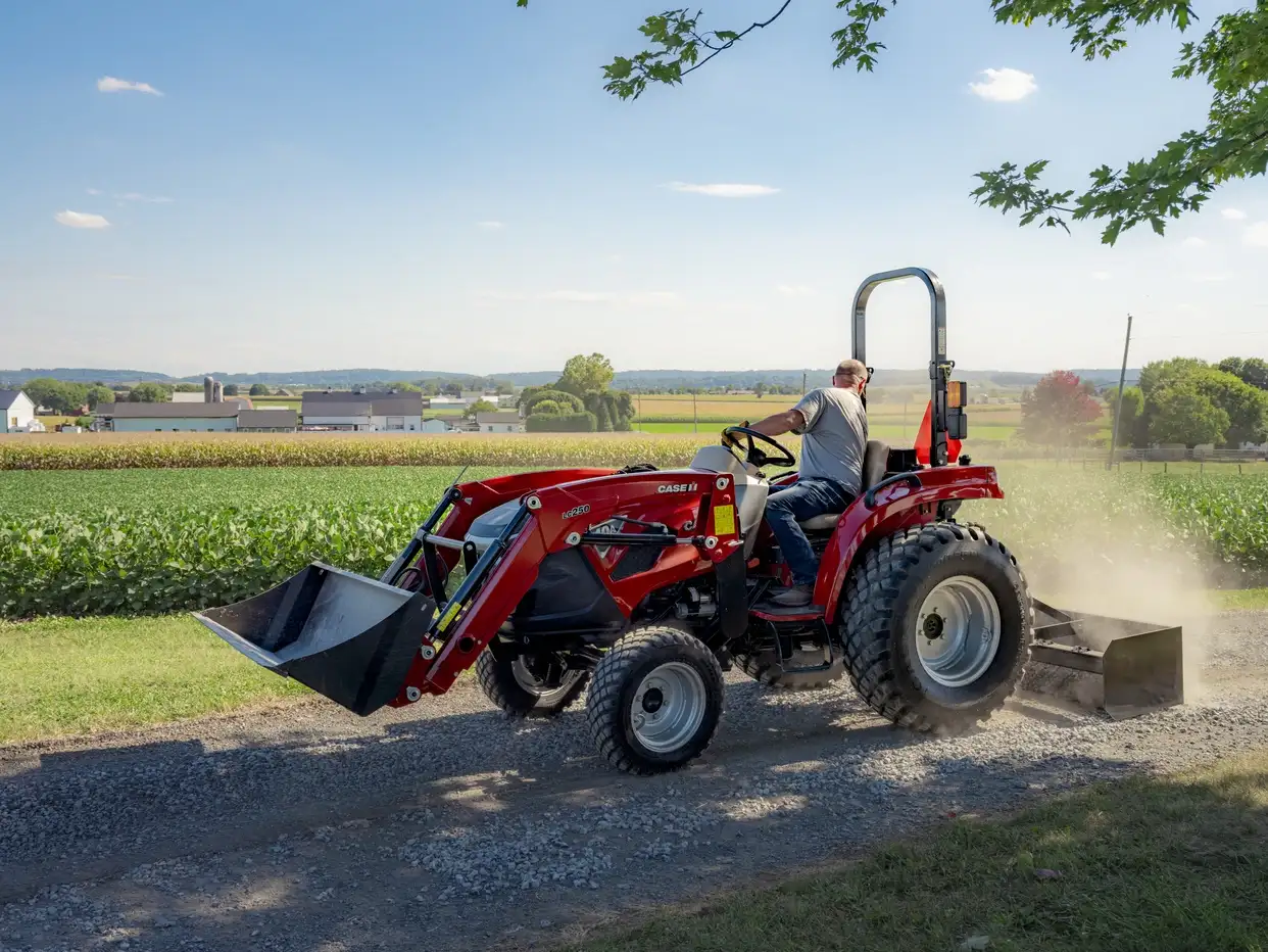 Red Case IH compact tractor with a farmer drives along a gravel path beside green fields, raising dust beneath a bright, sunny blue sky.