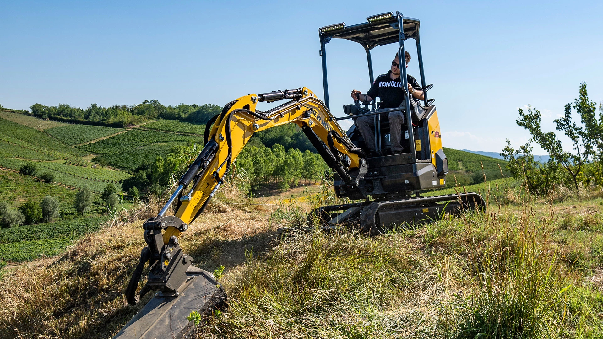 New Holland mini crawler excavator digging soil in agricultural field, village and trees in background.