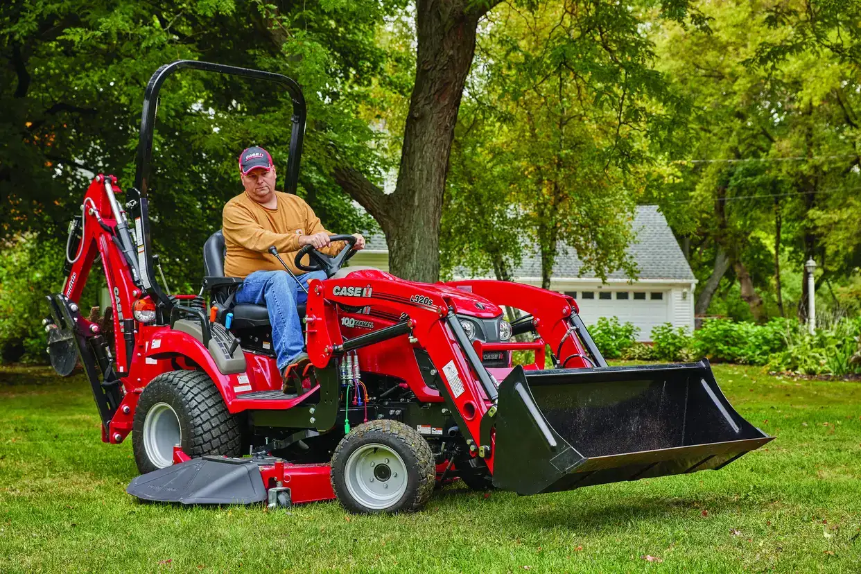 A person operates a Case IH subcompact utility tractor with a front loader in a grassy yard. 