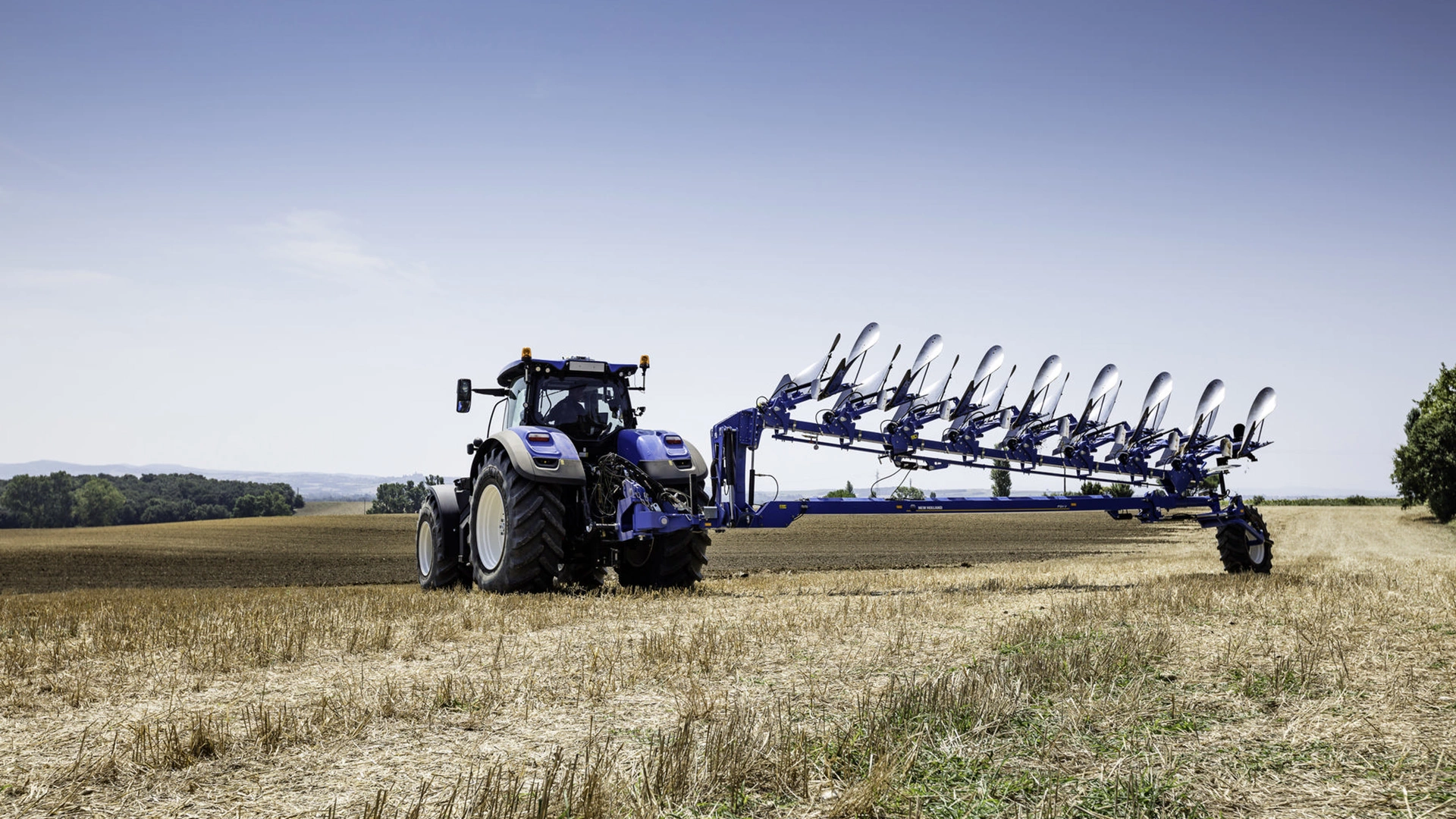 New Holland tractor with an extended reversible semi-mounted plough on a harvested field.