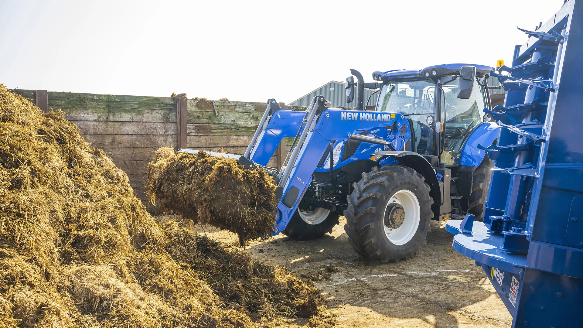New Holland blue tractor with front loader lifting manure at a farm.