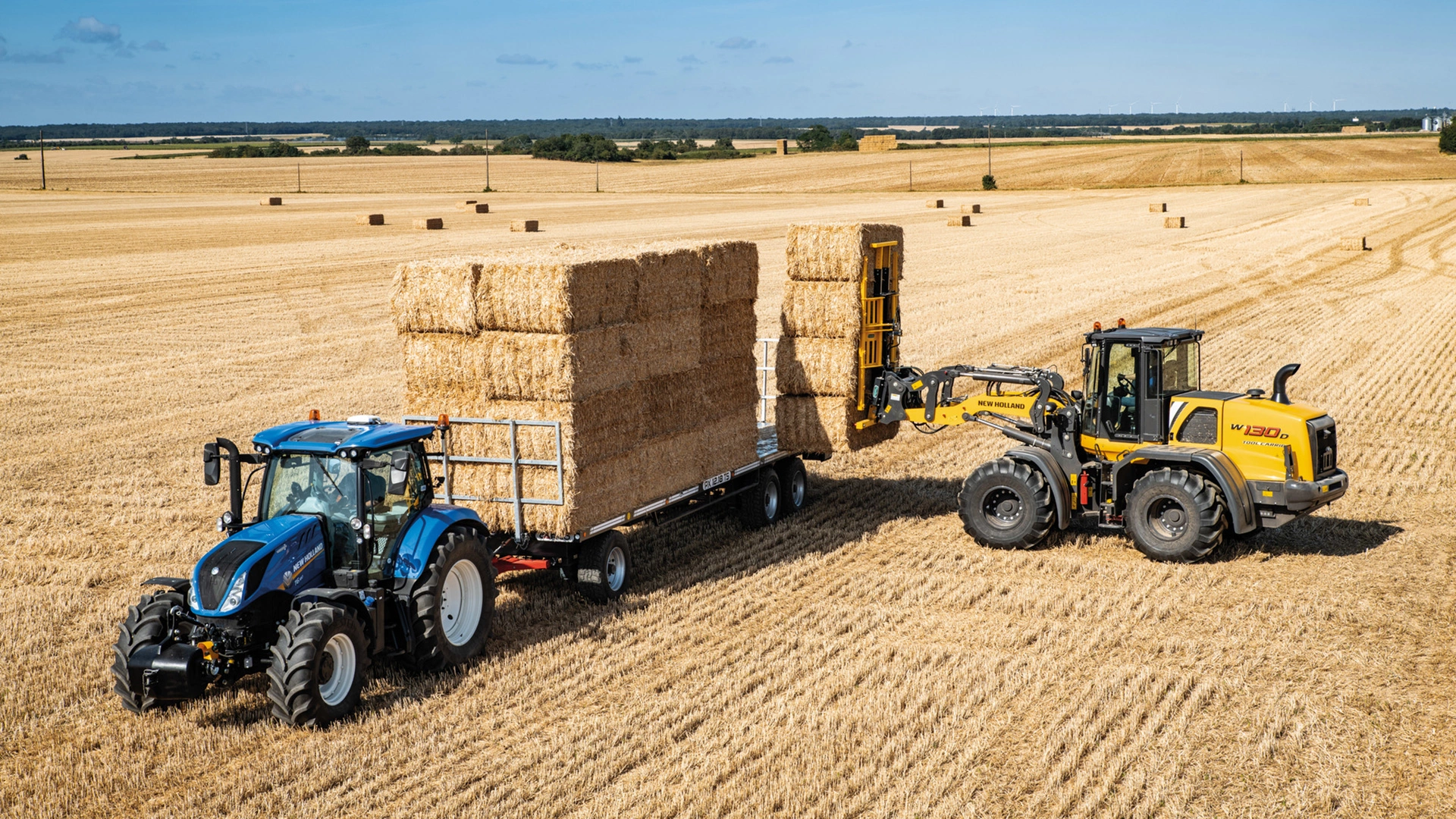 New Holland W300D wheel loader efficiently stacking large hay bales on a trailer in a harvested field.