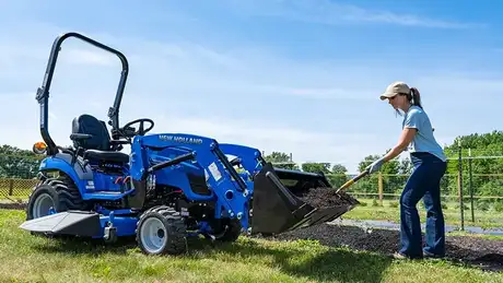 Woman with a shovel next to a blue tractor.