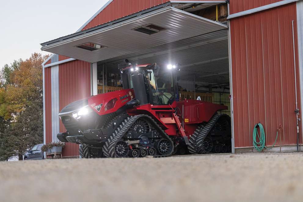 Red Case IH Steiger 785 tractor with rubber tracks parked inside a red barn with lights on during dusk.