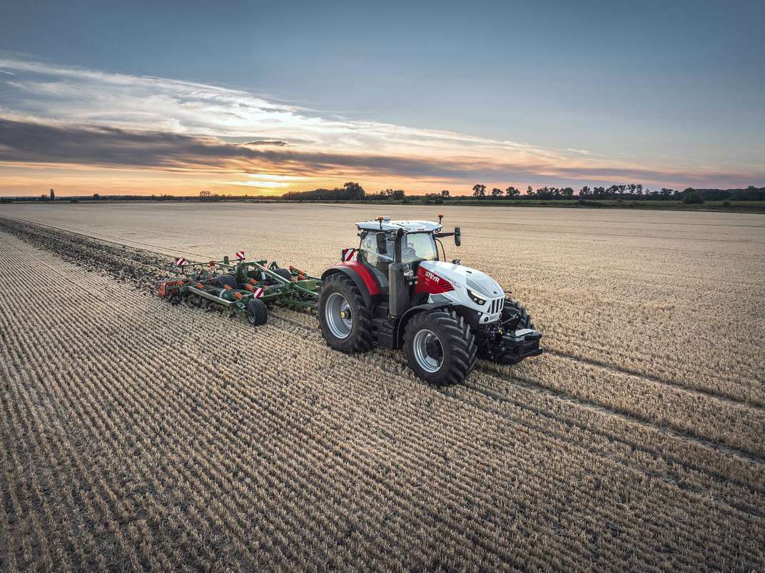 A white and red Steyr tractor pulling a green cultivator through a golden harvested field at sunset.
