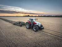 A white and red Steyr tractor pulling a green cultivator through a golden harvested field at sunset.