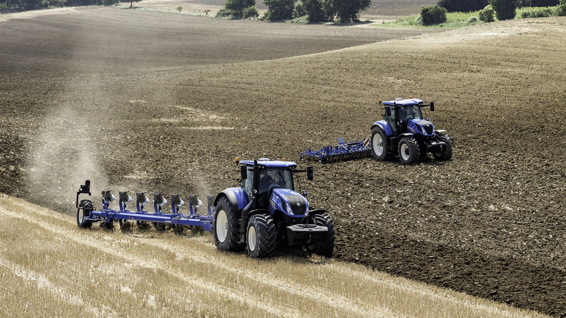 Two New Holland tractors in a vast field, one actively ploughing with a multi-furrow plough.