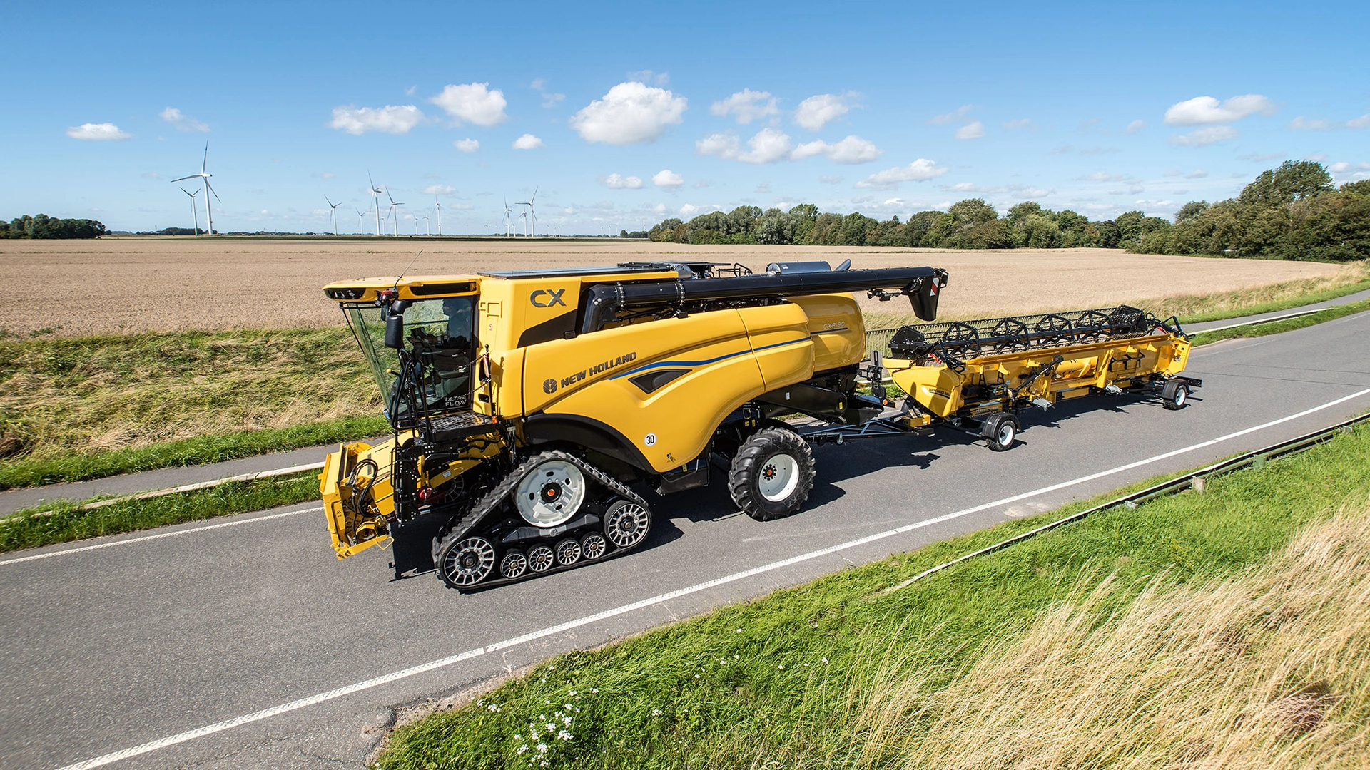 New Holland CX7 & CX8 Combine Harvesteron road, detached harvester equipment, fields & wind turbines background.