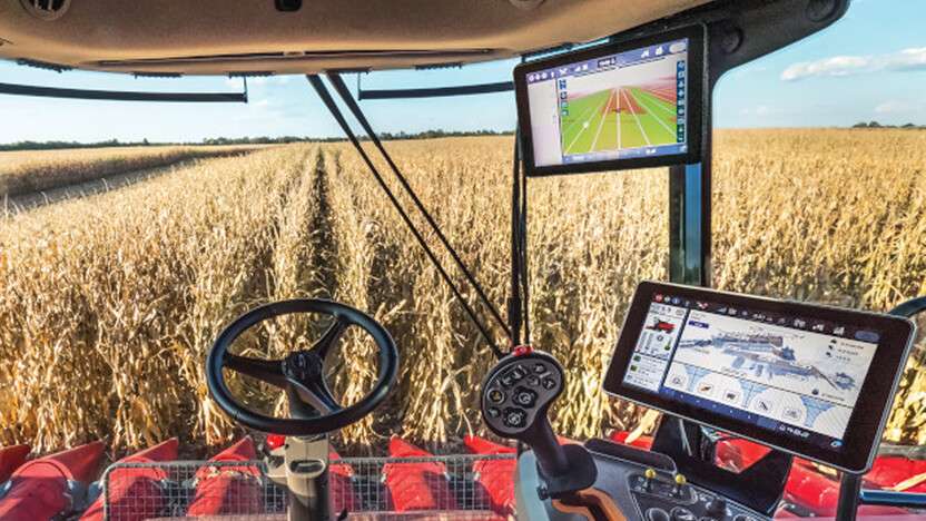 View from inside a Case IH tractor cab showing digital control screens and a steering wheel, overlooking a corn field being harvested.