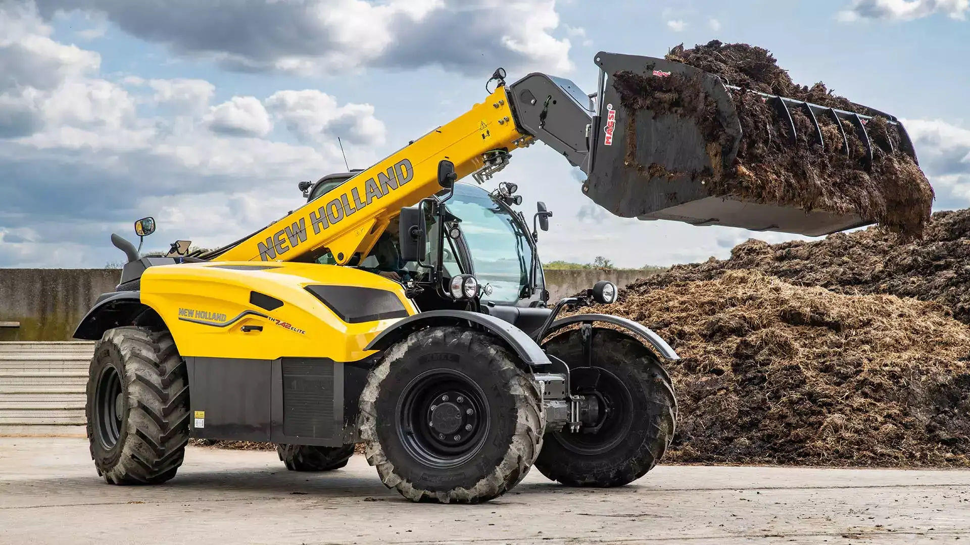 New Holland telehandler, model TH 7.42 Elite, lifting a full bucket of organic material on a farm.