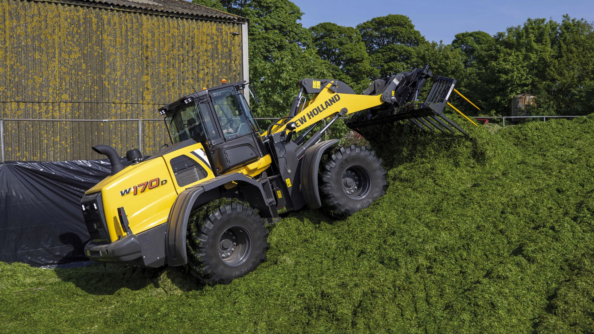 New Holland W170D wheel loader in action, lifting fresh green silage against a clear blue sky.