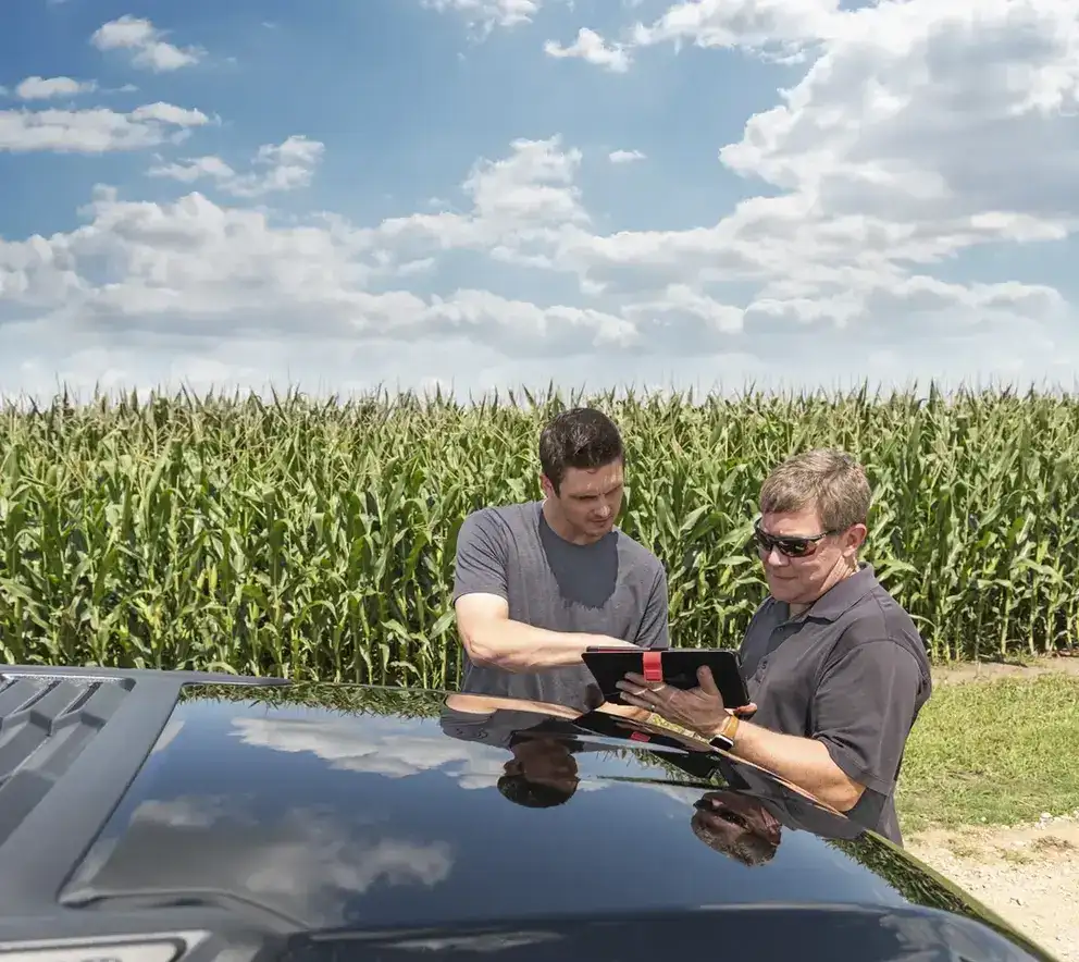 Two farmers review a tablet beside a vehicle in a tall cornfield under a bright, cloud-filled sky, conveying teamwork and rural productivity.
