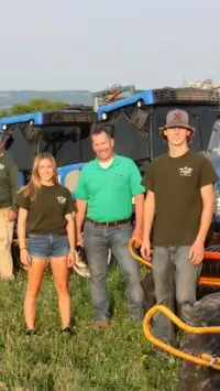 Family of 3 standing in front of New Holland tractors.