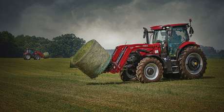 A Case IH tractor uses its front loader to lift a large round hay bale in a field, with another tractor and bale visible in the background.