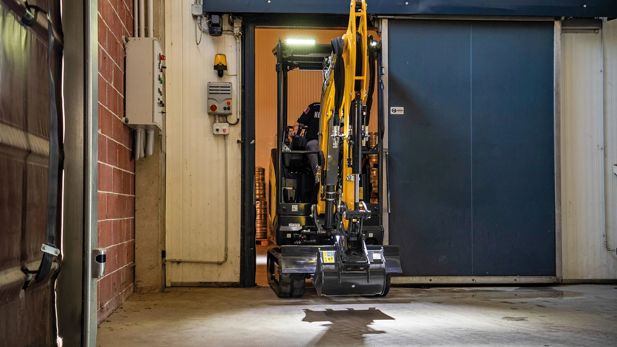 Compact New Holland mini excavator with an articulated arm and bucket exiting an industrial garage.