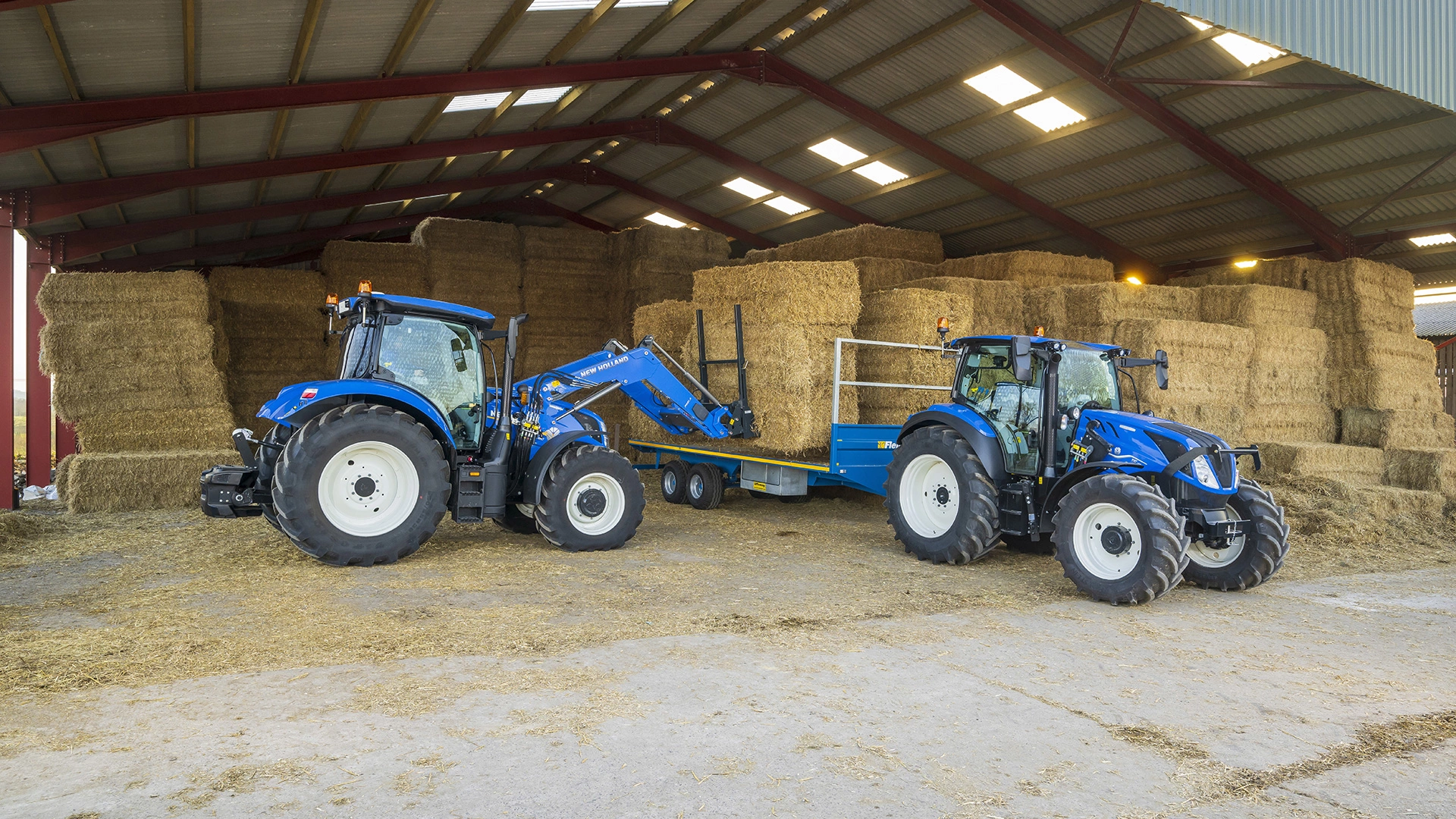 New Holland tractors in a barn, one with a front loader lifting straw bales onto a trailer.